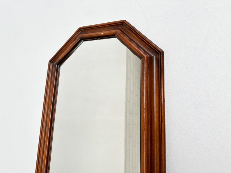 May include: A tall, octagonal mirror with a dark wood frame. The mirror is reflecting a white wall.