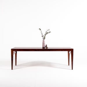 Può includere: Un lungo tavolo rettangolare in legno scuro con gambe affusolate. Un vaso con vegetazione e un piccolo oggetto decorativo sono appoggiati sulla superficie del tavolo. Il tavolo è lungo circa 183 cm.