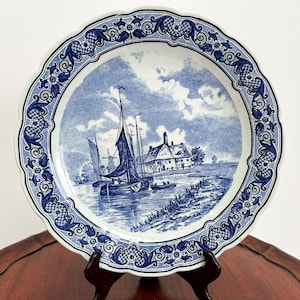 Puede incluir: Un plato de cerámica azul y blanco con una escena holandesa de una casa y barcos en un canal. El plato tiene un borde festoneado y un borde decorativo.