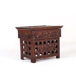 Puede incluir: Un cofre de madera marrón oscuro, tallado intrincadamente. La parte superior rectangular y la base cuadrada presentan tallas en relieve detalladas de figuras y patrones. El cofre se apoya sobre cuatro patas cortas. El diseño sugiere un estilo vintage o antiguo, de unos 60 cm de altura.