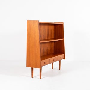 May include: A mid-century modern wooden bookcase with a warm, reddish-brown finish. The bookcase features two shelves, three small drawers, and a tall, angled back. The legs are slender and tapered.