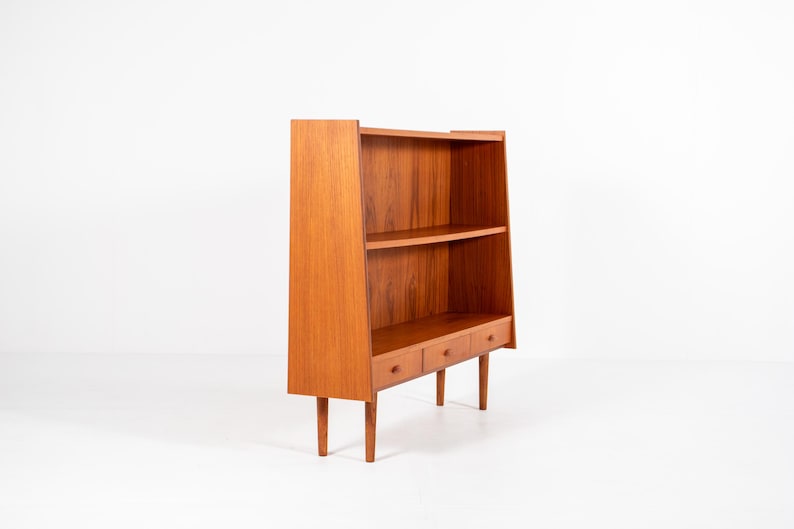 May include: A mid-century modern wooden bookcase with a warm, reddish-brown finish. The bookcase features two shelves, three small drawers, and a tall, angled back. The legs are slender and tapered.