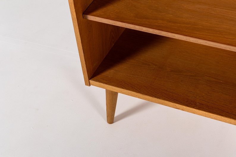 Puede incluir: Estanter&iacute;a de madera de estilo moderno de mediados de siglo, con dos estantes y patas c&oacute;nicas. La madera de color marr&oacute;n claro tiene un dise&ntilde;o sencillo y minimalista. Los estantes est&aacute;n soportados por un marco que se inclina ligeramente hacia afuera.
