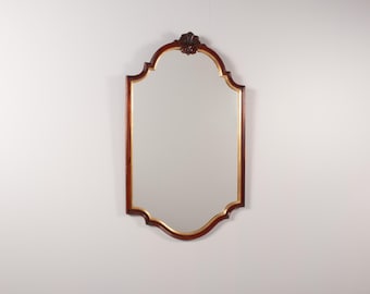 May include: A wall mirror with a dark brown wooden frame featuring a gold inner trim. The mirror has a scalloped, arched top and bottom edge with a decorative carved wooden detail centred at the very top of the frame.
