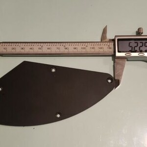 May include: A black, irregularly shaped guitar control cavity cover plate with three small screw holes. The plate is positioned against a metal digital caliper, which displays a measurement of 5.225 inches across its widest point.