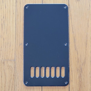 May include: A rectangular black plastic guitar tremolo cavity cover plate featuring six vertical oval-shaped string slots at the bottom and six small circular screw holes positioned around the perimeter of the flat, solid surface.