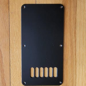 May include: A rectangular black plastic guitar tremolo cavity cover plate featuring six elongated oval string slots at the bottom and six small countersunk screw holes positioned around the perimeter for secure attachment to the instrument body.