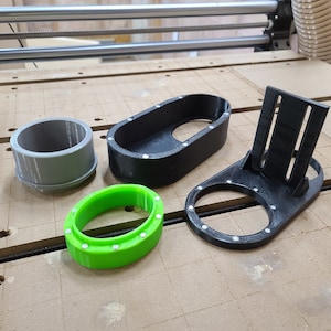 May include: Four 3D printed objects, including a black oval-shaped cup holder, a black stand with a cup holder, a gray cylinder, and a green ring.