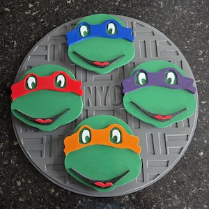 May include: Four Teenage Mutant Ninja Turtles face masks on a gray manhole cover. Each mask is green with a different colored eye mask: blue, red, purple, and orange.