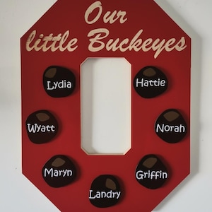 May include: A red wooden sign in the shape of the letter "O" with the words "Our little Buckeyes" in a light wood colour. The sign has seven dark brown, oval-shaped name tags with white lettering. Names include Lydia, Hattie, Wyatt, Norah, Maryn, Griffin, and Landry.