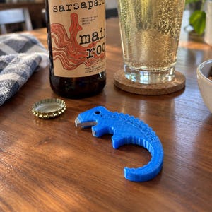 May include: A brown glass bottle of Main Root sarsaparilla with a tan label, a clear glass filled with a carbonated beverage, a gold metal bottle cap, and a blue plastic dinosaur-shaped bottle opener with a metal insert on a wooden surface.