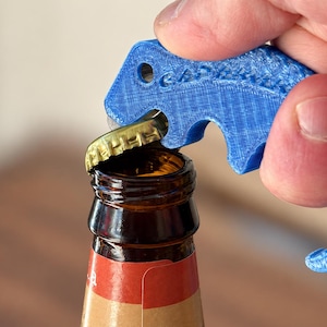Puede incluir: Un abridor de botellas azul impreso en 3D con forma de dinosaurio, con una superficie texturizada y el texto en relieve "OchPhant". El abridor está colocado sobre la chapa metálica dorada de una botella de vidrio marrón.