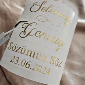 May include: A white adhesive transfer sheet with a grid pattern featuring metallic gold lettering that reads Selenay, Gencay, and S&ouml;z&uuml;m&uuml;z S&ouml;z, followed by the date 23.06.2024, with a small infinity symbol between the names and metal tweezers.
