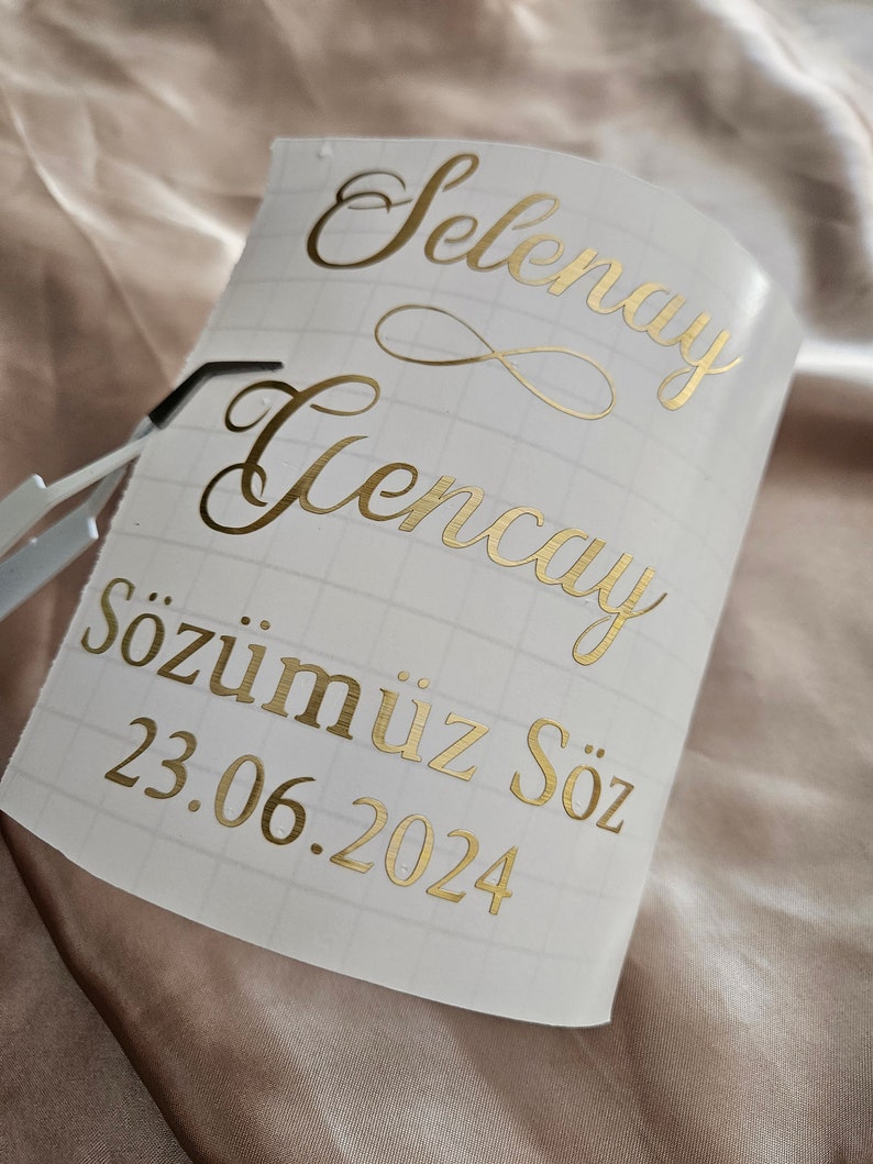 May include: A white adhesive transfer sheet with a grid pattern featuring metallic gold lettering that reads Selenay, Gencay, and S&ouml;z&uuml;m&uuml;z S&ouml;z, followed by the date 23.06.2024, with a small infinity symbol between the names and metal tweezers.