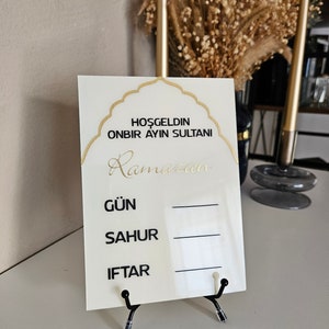 May include: A white sign with gold lettering that says "Hoşgeldin Onbir Ayın Sultanı Ramadan" and has a gold arch design. The sign has three blank lines with the words "Gün", "Sahur", and "Iftar" written in black.