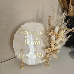 Könnte beinhalten: Ein rundes, klares Acrylschild mit goldener Beschriftung, auf dem "Sozlumuz So Sevda & Enes" steht. Das Schild ist mit Trockenblumen und einem goldenen Band verziert und steht auf einem goldenen Metallständer.