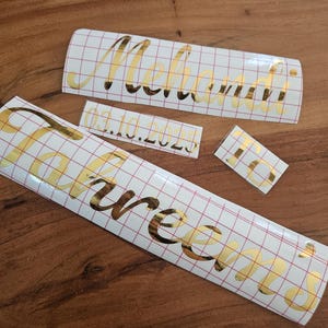 May include: Three gold metallic vinyl decals with a cursive font on a white grid transfer paper. The decals feature the names Mehandi and Tahreem, the date 03.10.2025, and the letters to. The adhesive lettering is designed for application to surfaces.