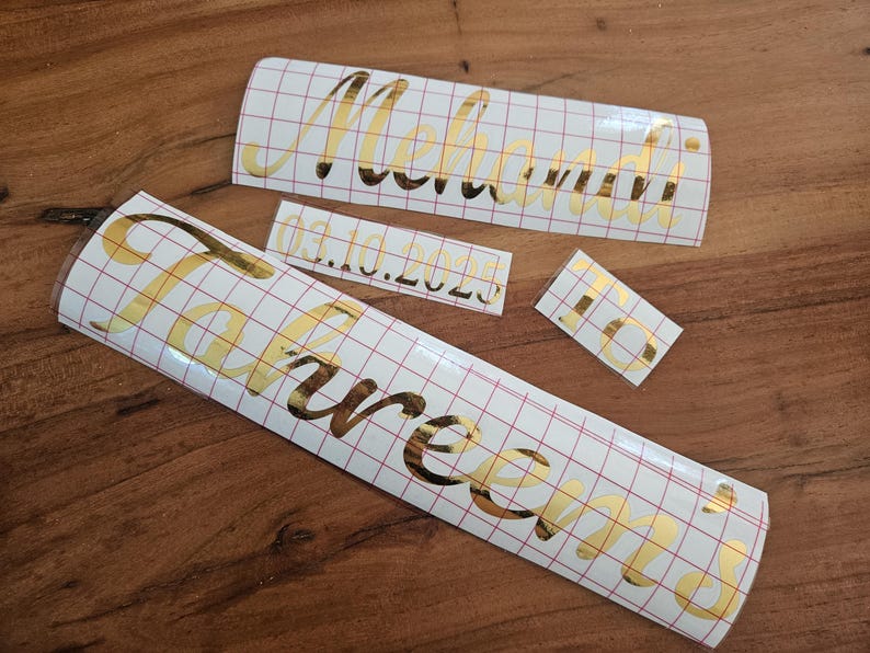 May include: Three gold metallic vinyl decals with a cursive font on a white grid transfer paper. The decals feature the names Mehandi and Tahreem, the date 03.10.2025, and the letters to. The adhesive lettering is designed for application to surfaces.