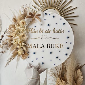 Könnte beinhalten: Ein rundes weißes Schild mit dem goldenen Text Hûn bi xêr hatin und MALA BUKE sowie einem Herzsymbol. Es ist mit getrockneten beigen Blumen, Gräsern und kleinen blauen Nazar-Perlen verziert und mit einer braunen Schleife versehen.
