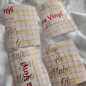 May include: Four rectangular sheets of gold metallic vinyl decals featuring the text "Bajram Serif Mubarek Olsun" in a cursive script. Each sheet is covered with a clear transfer tape marked with a red grid and the text "Cricut Joy Vinyl".