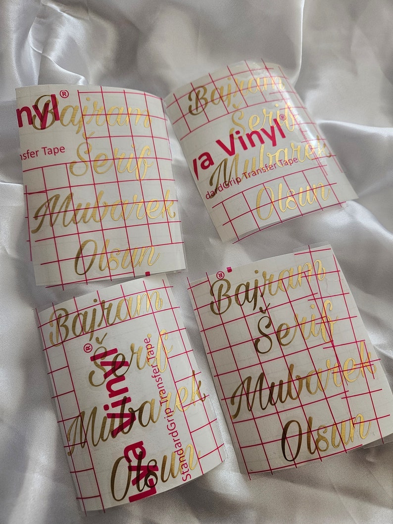 May include: Four rectangular sheets of gold metallic vinyl decals featuring the text "Bajram Serif Mubarek Olsun" in a cursive script. Each sheet is covered with a clear transfer tape marked with a red grid and the text "Cricut Joy Vinyl".