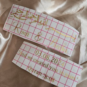 May include: Two rectangular adhesive vinyl decals with gold lettering on a white grid transfer paper. The top decal features Arabic calligraphy and the names Rima & Bechir. The bottom decal displays the date 03.08.2024 and the text And we created you in pairs. Quran 78:08.