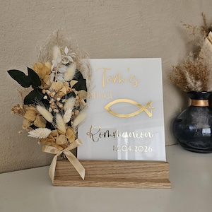 Könnte beinhalten: Ein weißes Acrylschild mit goldenen Buchstaben und einem goldenen Fischsymbol, das auf einem Holzsockel steht. Auf dem Schild steht "Tom's", "Heilige", "Kommunion" und "12.04.2026". Ein Strauß aus Trockenblumen und Grünzeug ist mit einem beigen Band gebunden.