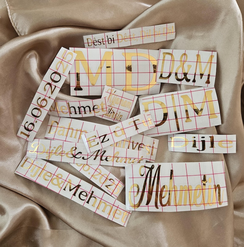 May include: A collection of gold foil adhesive vinyl decals featuring various names, dates, and phrases including D&egrave;st bi D&ecirc;st, Dil bi Dil, MD, D&M, 16.06.2025, Mehmet, Ez &ucirc; Tu, Dijle, and Dijle & Mehmet, all presented on white transfer paper with a red grid.