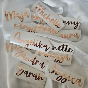 May include: A collection of custom vinyl name decals in a metallic rose gold finish, featuring various names such as Maja, Michelle, Angelika, and Jessica written in a cursive script font, each applied to a white rectangular backing paper.