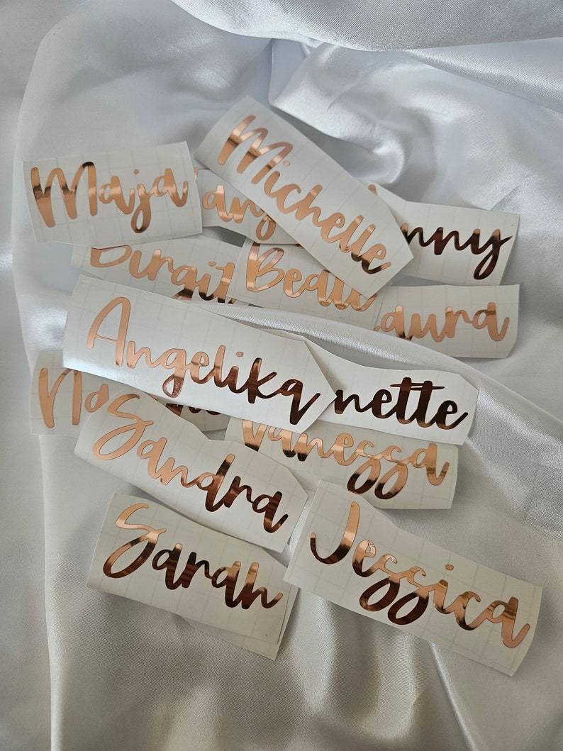 May include: A collection of custom vinyl name decals in a metallic rose gold finish, featuring various names such as Maja, Michelle, Angelika, and Jessica written in a cursive script font, each applied to a white rectangular backing paper.