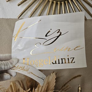 May include: A white adhesive transfer sheet featuring gold metallic lettering that reads "Kiz Evi" and "Hosgeldiniz" in a mix of script and serif fonts, with a smaller separate strip below displaying Arabic calligraphy in gold.