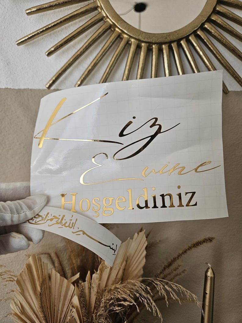 May include: A white adhesive transfer sheet featuring gold metallic lettering that reads "Kiz Evi" and "Hosgeldiniz" in a mix of script and serif fonts, with a smaller separate strip below displaying Arabic calligraphy in gold.