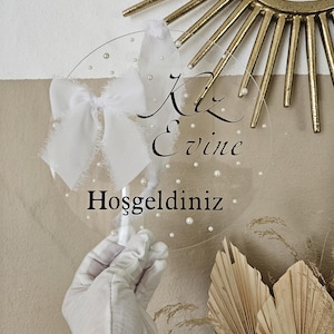 May include: A clear round acrylic sign with white pearl accents and a white satin bow. The sign says "Kiz Evine Hosgeldiniz" in black cursive lettering.