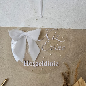 May include: A clear round acrylic sign with a white bow and the text "Kiz Ervine" and "Hosgeldiniz" in white lettering.