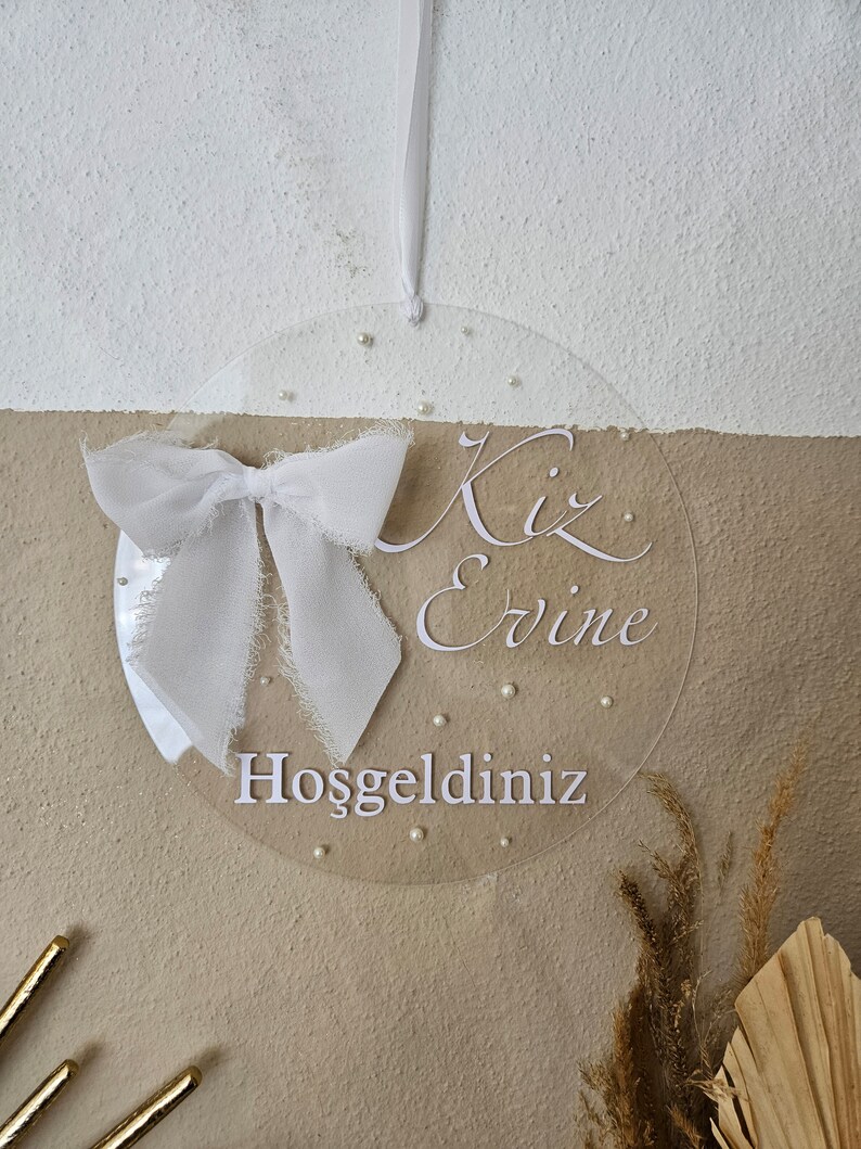 May include: A clear round acrylic sign with a white bow and the text "Kiz Ervine" and "Hosgeldiniz" in white lettering.