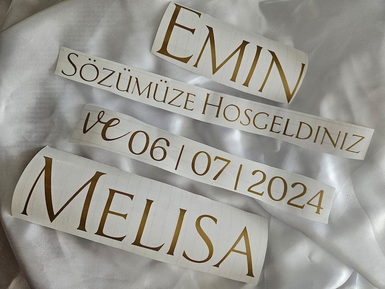 May include: Four separate adhesive vinyl decals featuring gold lettering on a white backing. The text reads EMIN, S&Ouml;Z&Uuml;M&Uuml;ZE HOSGELDINIZ, ve 06 | 07 | 2024, and MELISA. The decals are arranged in a stack, showcasing a mix of serif and script fonts.
