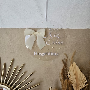 May include: A clear round sign with a white ribbon bow and the text "Kiz Evine Hosgeldiniz" which translates to "Welcome to the Girl's Home" in English.