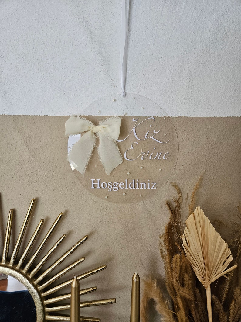 May include: A clear round sign with a white ribbon bow and the text "Kiz Evine Hosgeldiniz" which translates to "Welcome to the Girl's Home" in English.