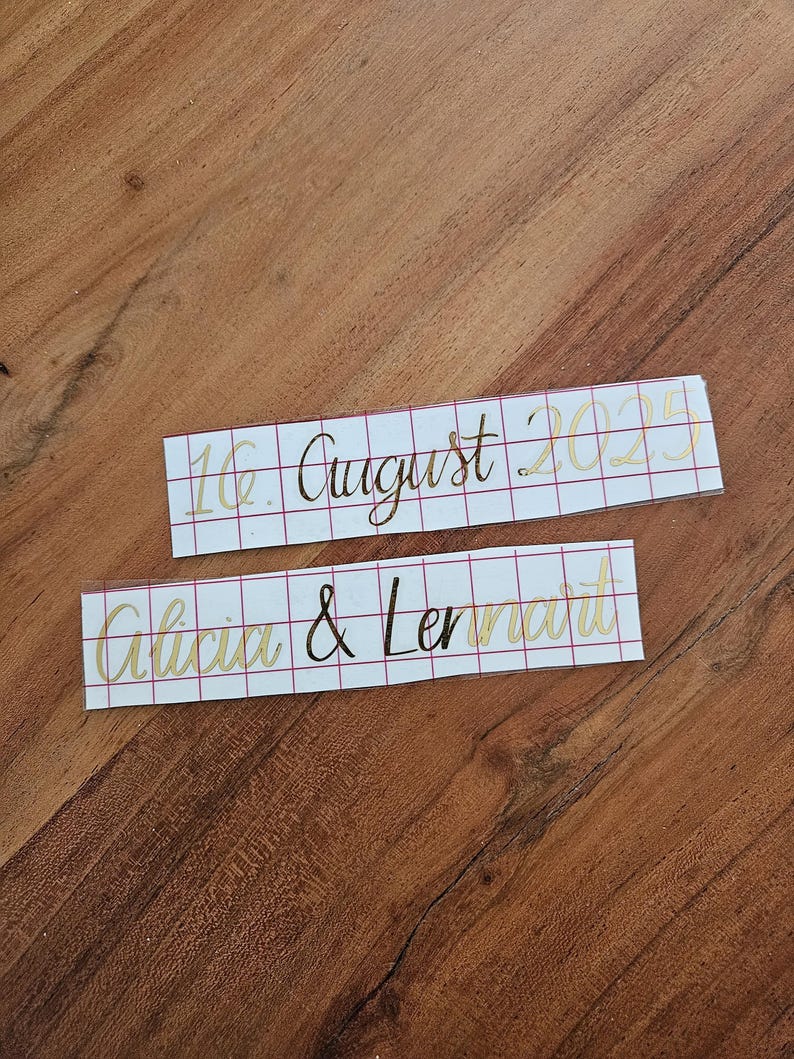 May include: Two rectangular adhesive vinyl decals with a red grid transfer paper backing. The top decal features the gold text 16. August 2025. The bottom decal displays the gold names Alicia & Lennart in a cursive script font style.
