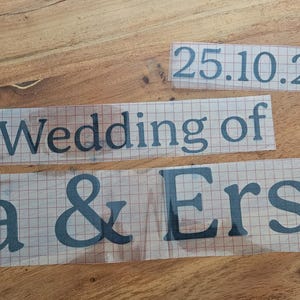 May include: Three black vinyl adhesive decals on a clear transfer sheet with a grid pattern. The text reads 25.10.2025, The Wedding of, and Lea & Ersan in a serif font. These custom lettering stickers are designed for application onto smooth surfaces.