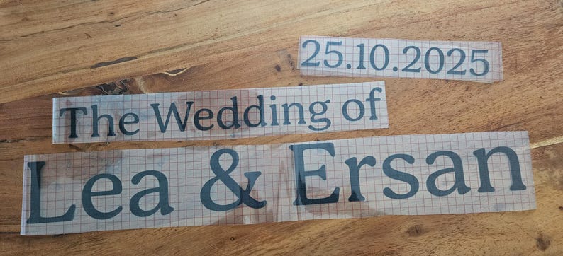 May include: Three black vinyl adhesive decals on a clear transfer sheet with a grid pattern. The text reads 25.10.2025, The Wedding of, and Lea & Ersan in a serif font. These custom lettering stickers are designed for application onto smooth surfaces.