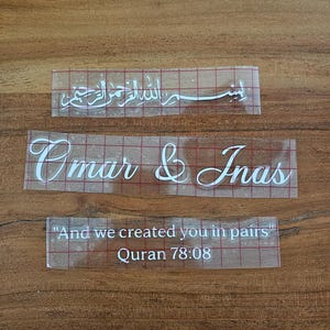 May include: Three white vinyl adhesive decals on clear transfer tape with a red grid pattern. The top decal features Arabic calligraphy, the middle decal reads "Omar & Inas" in script, and the bottom decal reads "And we created you in pairs" Quran 78:08.