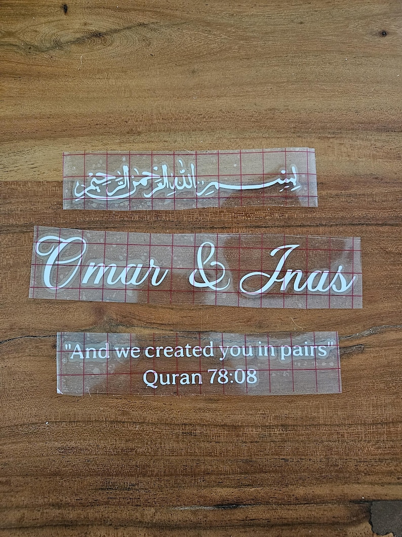 May include: Three white vinyl adhesive decals on clear transfer tape with a red grid pattern. The top decal features Arabic calligraphy, the middle decal reads "Omar & Inas" in script, and the bottom decal reads "And we created you in pairs" Quran 78:08.