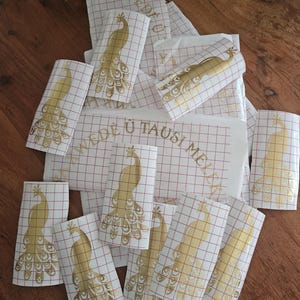 May include: A collection of gold peacock vinyl decals on white transfer paper with a red grid pattern. Several individual peacock stickers are visible, along with a larger rectangular sheet featuring the text YAWEDE U TAUSI MELEK.