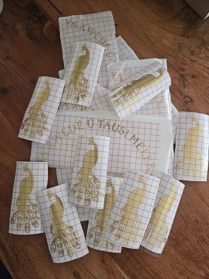 May include: A collection of gold peacock vinyl decals on white transfer paper with a red grid pattern. Several individual peacock stickers are visible, along with a larger rectangular sheet featuring the text YAWEDE U TAUSI MELEK.