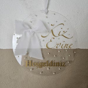 May include: A clear round acrylic sign with a white bow and gold lettering that reads "Kiz Evine Hosgeldiniz".