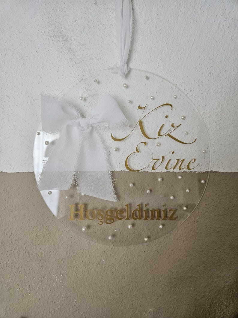 May include: A clear round acrylic sign with a white bow and gold lettering that reads "Kiz Evine Hosgeldiniz".