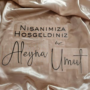 May include: Black vinyl lettering decals on a grid transfer paper, featuring the text NISANIMIZA HOSGELDINIZ, ve, Aleyna, and Umut, placed on a smooth, champagne-colored satin fabric with soft folds and a reflective, glossy finish.