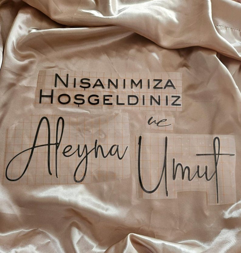 May include: Black vinyl lettering decals on a grid transfer paper, featuring the text NISANIMIZA HOSGELDINIZ, ve, Aleyna, and Umut, placed on a smooth, champagne-colored satin fabric with soft folds and a reflective, glossy finish.