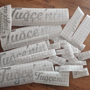 May include: A collection of silver vinyl decals on white transfer paper with a red grid pattern. The decals feature the text Tuğ&ccedil;e'nin and NİŞAN BOH&Ccedil;ASI in a cursive script font, designed for application onto various surfaces as custom labels.